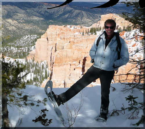 Photo of Jerry Michelsen in snoe shoes with one foot raised. It's Bryce Canyon National Park, in snow, overlooking a beautiful gorge with mountains in the distance.